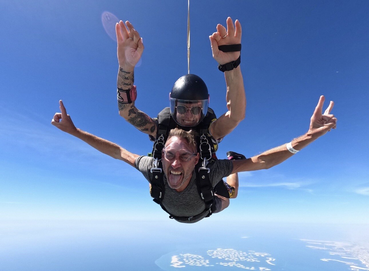 Jesse Judelman tandem skydiving above the coastline under a clear blue sky.